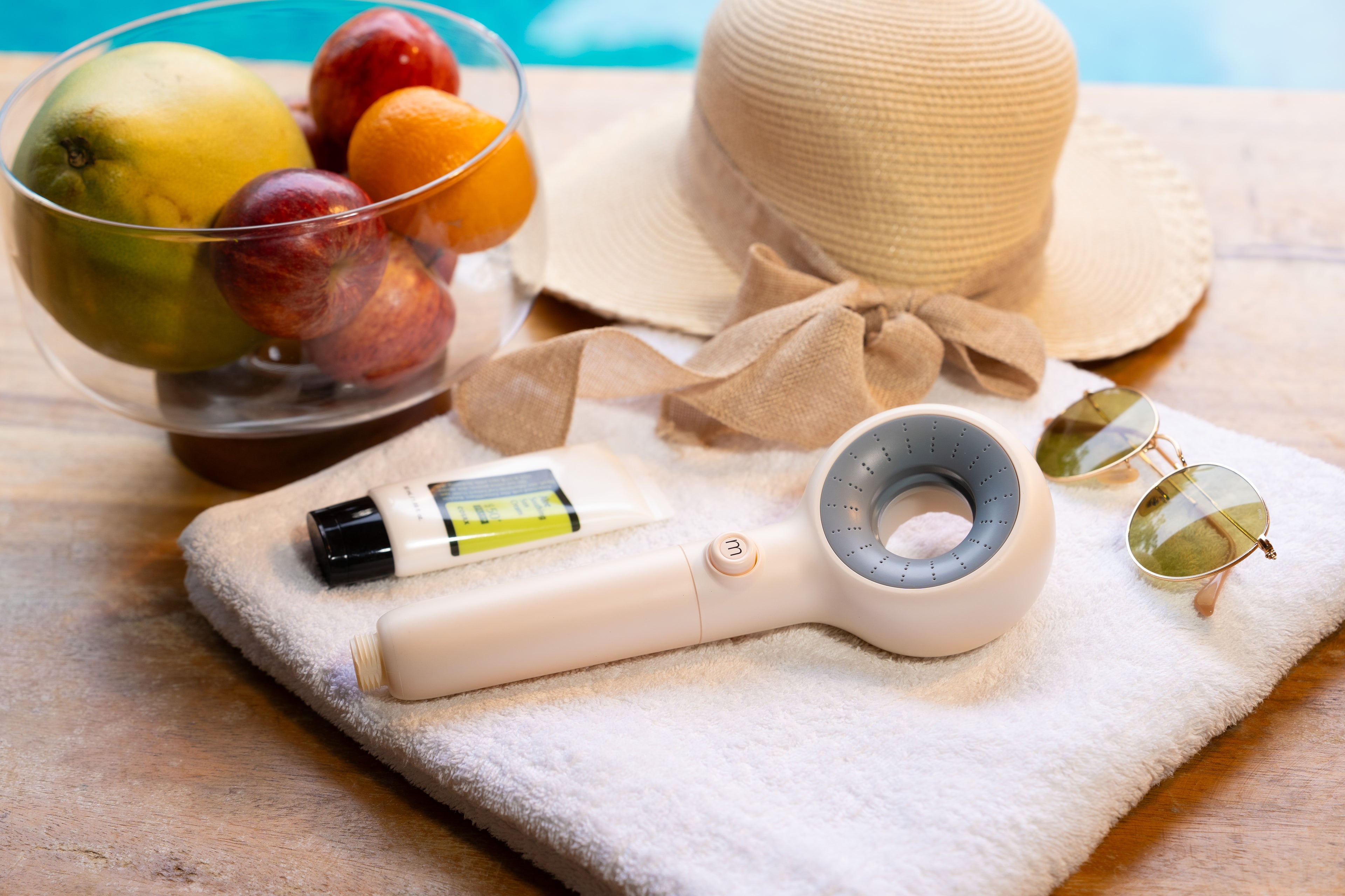Personal care items including a tube of cream, Murelle shower head and sunglasses on a towel with fruits and a hat in the background.