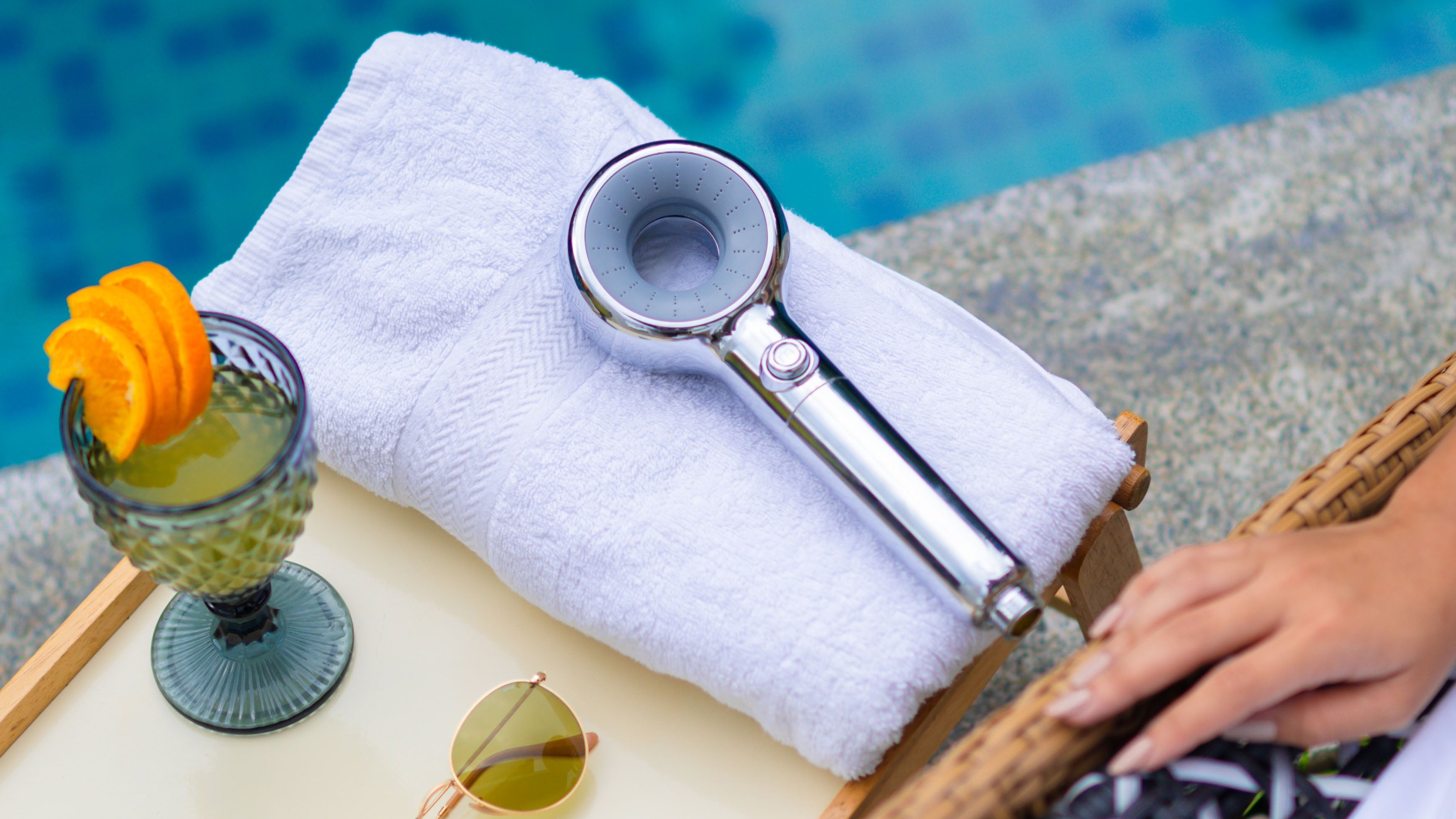 Cocktail with a slice of mango, sunglasses, and a handheld showerhead on a towel by a pool.